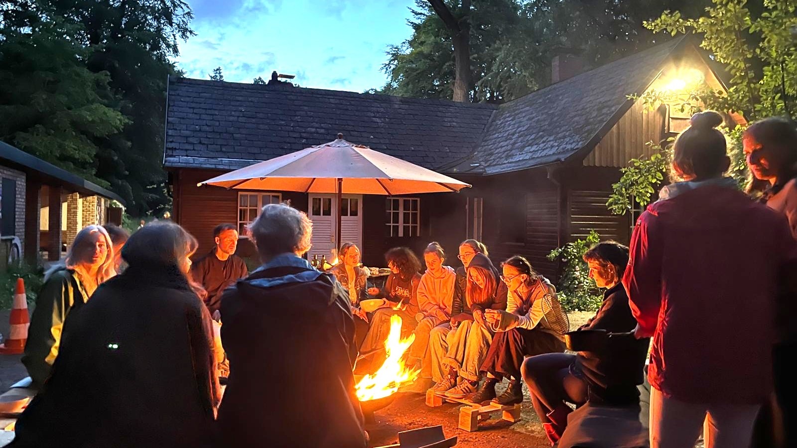 Menschen sitzen abends um ein Feuer vor dem Waldhaus im Heilgarten Hamburg